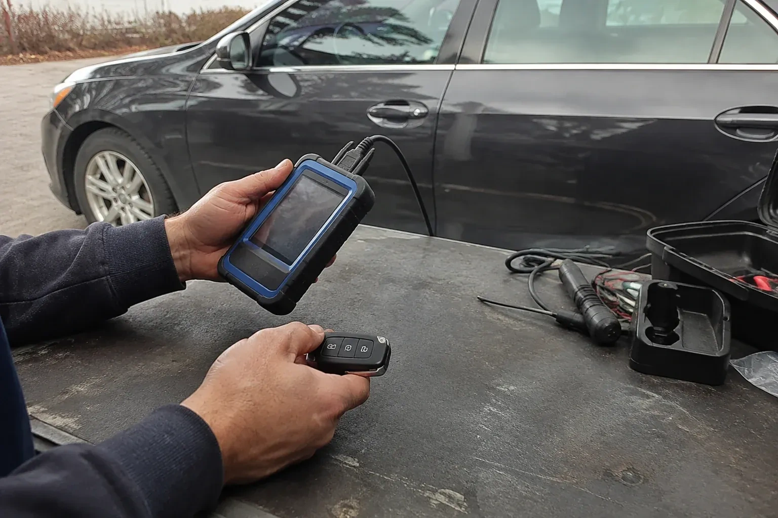 Hands holding a blue-edged electronic device and car-key fob at a worktable beside a parked car. — Transponder Key Programming, Woolwich