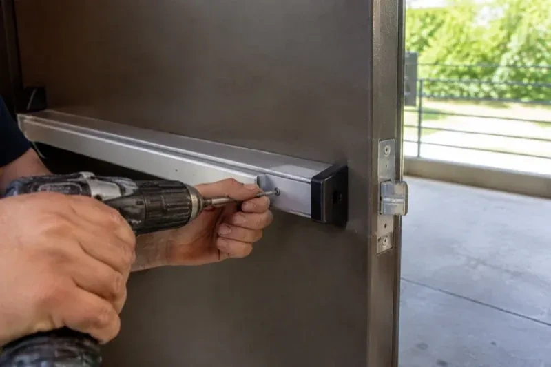 Hands using a power driver at the end of a horizontal push bar on an open metal door. — Panic & Exit Hardware, Woolwich